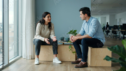 Two professional colleagues having a conversational meeting while sitting on wooden blocks in modern office with engaged conversation natural light streaming large window