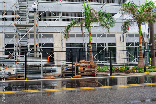 Scaffolding and construction materials line a downtown building frontage flanked by palm trees, wet pavement and reflective puddles after rain, conveying urban renovation and temporary disruption.
