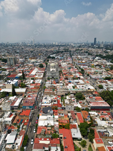 Stunning vertical drone panorama capturing the Holy Family Parish Church and the dense urban landscape of Guadalajara, Mexico. Modern buildings rise in the distance under a partly cloudy sky, showcasi