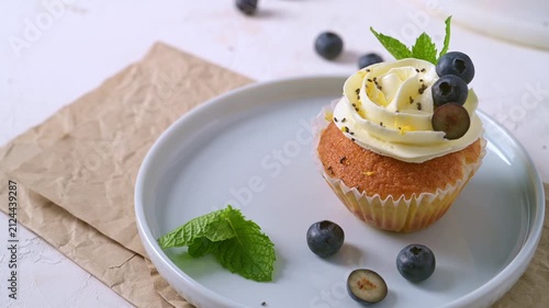 A freshly baked vanilla cupcake with rich white cream frosting is decorated with fresh blueberries, chia seeds, and mint leaves under bright natural light
