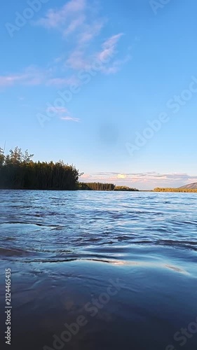 Serene lake with gentle waves reflecting a clear blue sky, bordered by dense green forest. Peaceful natural landscape ideal for relaxation.