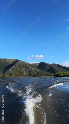 View from a boat on a river creating a wake, surrounded by lush green mountains under a bright blue sky with scattered clouds.