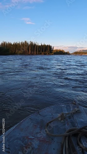 A small boat floats on calm water under a clear blue sky at sunset, featuring a forested shoreline and ropes on its deck.