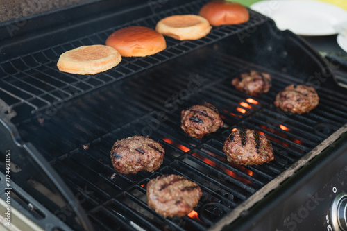 Several homemade beef burger patties cook over glowing flames on a gas grill while brioche buns toast on the upper warming rack. This image highlights backyard barbecues, and outdoor cooking