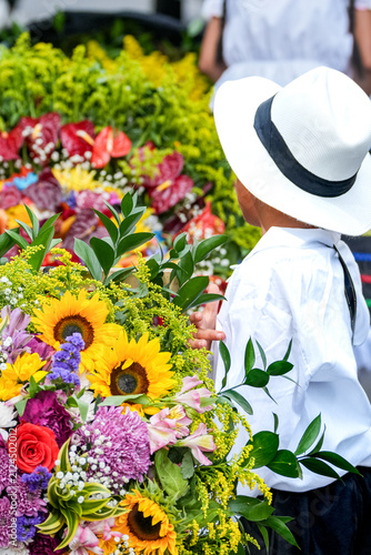 Young boy wearing traditional Colombian clothes and white hat carrying a vibrant silleta flower arrangement during the Flower Festival parade in Medellin Colombia
