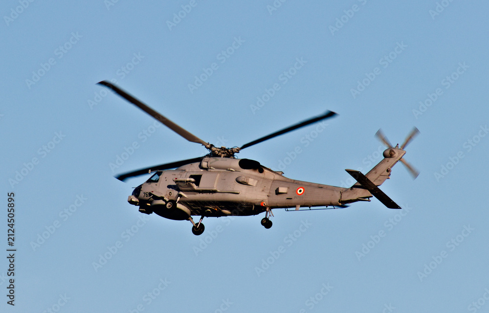 Fototapeta premium A naval military helicopter flies through a clear blue sky with landing gear lowered during an apparent coastal aviation operation.
