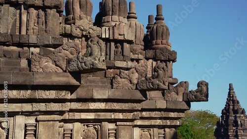 Carved temple wall leads toward a distant Prambanan spire under brilliant tropical sunlight and cloudless blue sky.