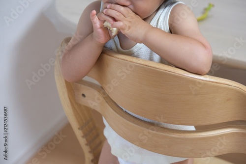 Close-up of a toddler eating a banana with both hands on a wooden chair
