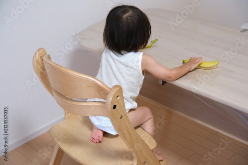Back view of a toddler wearing a diaper sitting on a wooden chair reaching for a banana peel on the table