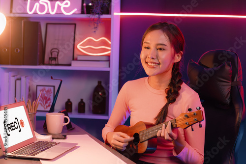Foto Host channel in musician of young beautiful Asian playing ukulele along singing, broadcasting on social media channel by phone at neon colorful light modern studio recording by camera