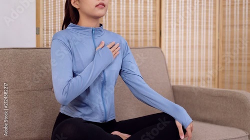 Young Asian woman holding chest experiencing breathing difficulty during yoga exercise at home