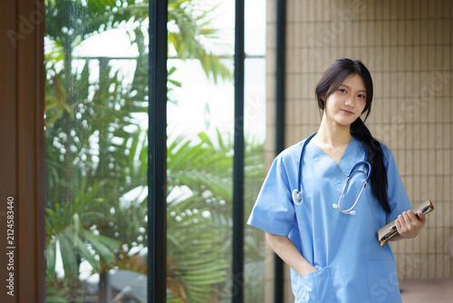 Confident young female nurse in blue scrubs stands in a modern hospital corridor, holding a clipboard and smiling warmly. Professional healthcare worker ready to assist patients