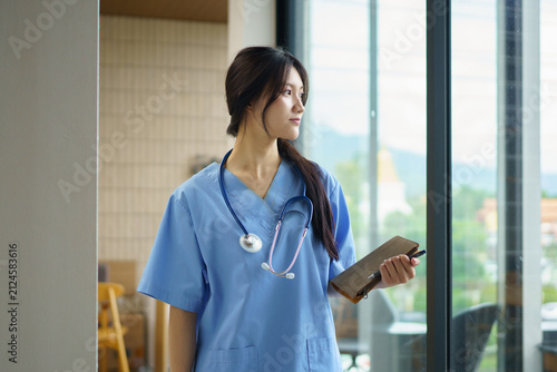 Confident young female nurse in blue scrubs stands in a modern hospital corridor, holding a clipboard and smiling warmly. Professional healthcare worker ready to assist patients