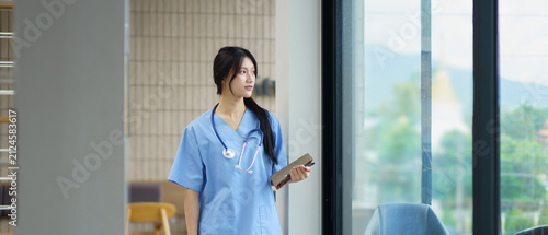 Confident young female nurse in blue scrubs stands in a modern hospital corridor, holding a clipboard and smiling warmly. Professional healthcare worker ready to assist patients