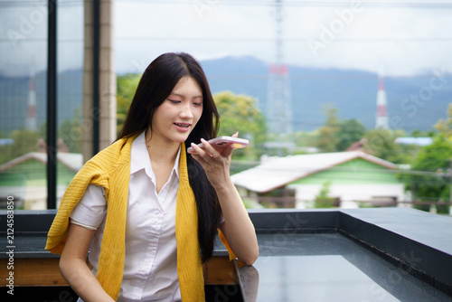 A young Asian woman speaks into her smartphone using voice message feature at Cafe with Mountain View Background, conveying casual communication and technology lifestyle