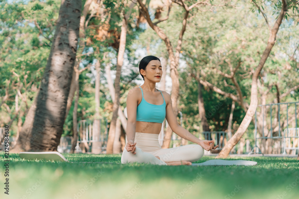 Fototapeta premium Asian woman meditating in a peaceful park setting, sitting cross-legged on green grass with trees in the background on a sunny day. Concept of mindfulness, self-care, and outdoor relaxation
