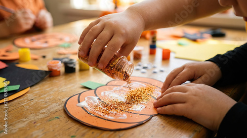 Child hands pouring glitter on paper pumpkin craft with glue on wooden table, autumn art activity for kids, creative and fun seasonal project