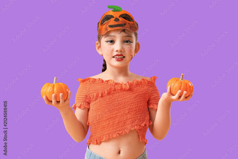 Obraz premium Young girl in pumpkin mask and orange top holding two small pumpkins for halloween celebration, She has playful face paint and a braided hairstyle