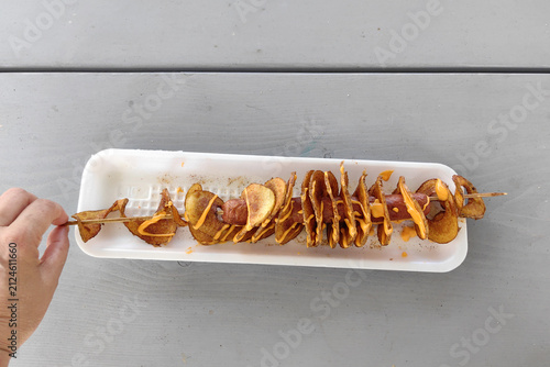 Картината върху платно A hand picks up a skewer with a baked sausage and fried spiral potato, resting on a serving tray