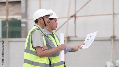 Two Civil Engineers in Hard Hats and Safety Vests Inspecting Construction Site with Blueprint