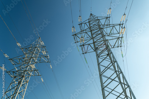 High voltage transmission towers and power lines stretching across clear blue sky in low angle industrial infrastructure scene with strong sunlight and extensive overhead network