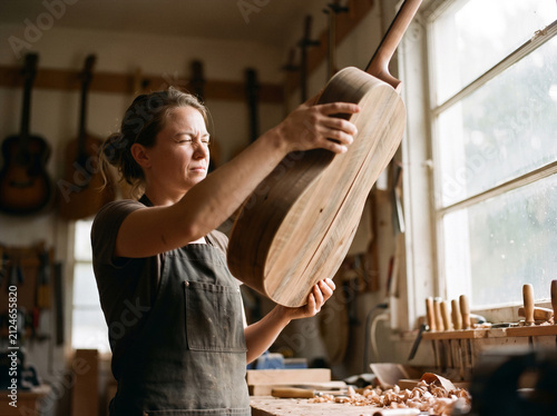 Female luthier inspecting handmade wooden acoustic guitar finish by window light, craftsman woodworking art and music instrument manufacturing lifestyle concept