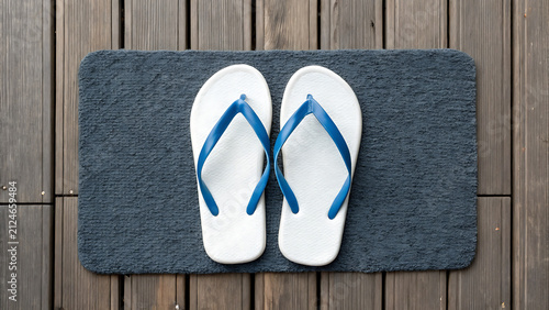 White flip flops on gray doormat at wooden entryway. Casual summer footwear placed outdoors for beach or vacation styling.