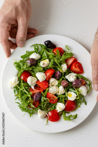 Healthy summer salad with mozzarella cheese, tomatoes, arugula and olives on white plate serving, olive oil garnish. Top view