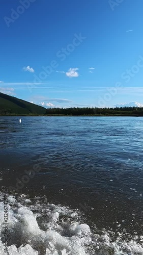 Gentle waves lapping at the shore with foamy water, set against a clear blue sky and distant forested hills. Serene natural landscape featuring calm waters, scattered clouds, and mountainous backdrop.