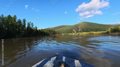 Calm river from a boat's perspective, surrounded by dense forests and lush green hills under a bright blue sky with scattered clouds.
