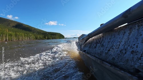 Boat cruising on a river with white wake, surrounded by dense forests and mountains under clear blue sky.