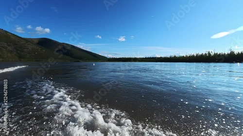 Scenic view of a river with gentle waves reflecting sunlight, bordered by lush green hills and dense forests under a bright blue sky with scattered clouds.