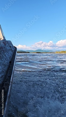 View from a moving boat on a river, showing water splashes and ripples. Background features clear blue sky with scattered clouds, distant mountains, and shoreline vegetation.