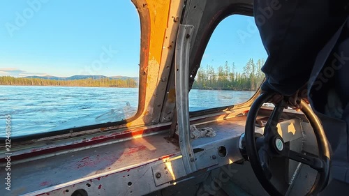View from inside a weathered boat showing rusted metal frames, water, and forested shore under sunlight. Driver in high-visibility jacket steering.