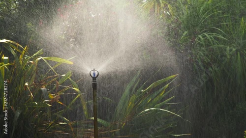 A tall commercial-grade sprinkler sprays a heavy volume of water across a lush, healthy lawn in a large garden under bright summer sunlight, surrounded by dense green foliage.