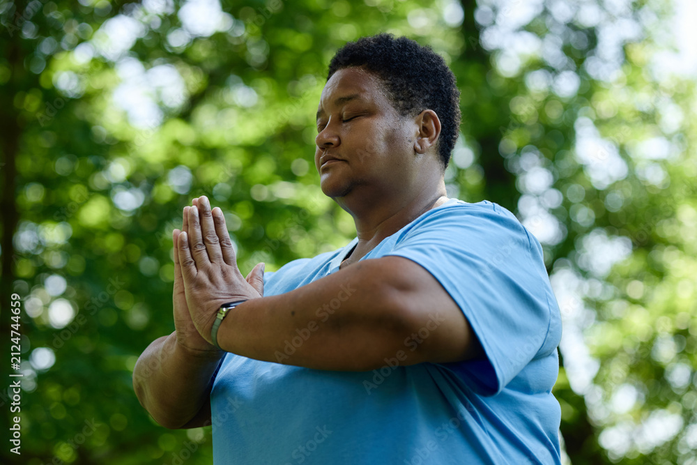Naklejka premium Middle aged African American woman meditating with closed eyes and hands in prayer position in park, promoting mental wellbeing and outdoor wellness campaigns, medium shot