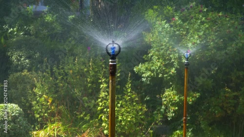 A tall commercial-grade sprinkler sprays a heavy volume of water across a lush, healthy lawn in a large garden under bright summer sunlight, surrounded by dense green foliage.