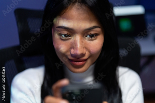 Принти на полотні Close up of a young woman smiling as she looks down at her smartphone in a dark room lit by blue and green screen glow, absorbed in a mobile app at night