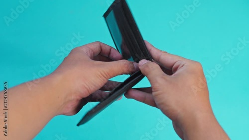 Hands open empty black wallet letting remaining coins fall out onto turquoise background