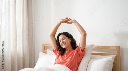 Happy mid-adult woman stretching and waking up in a bright bedroom.