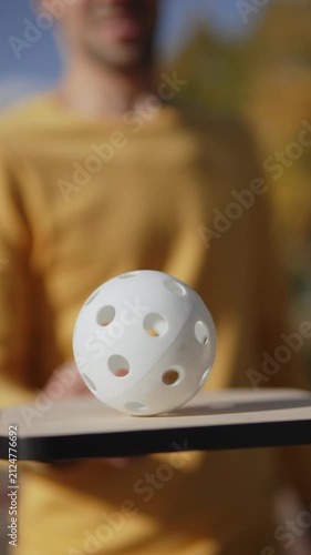 Pickleballer rolling perforated white ball on wooden paddle, vertical video. Balance and coordination training, preparation for pickleball match in park on outdoor arena.
