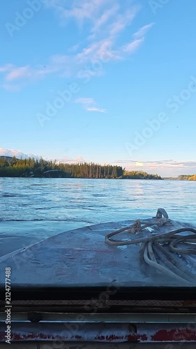 Serene view from a boat on calm river waters, surrounded by dense green forests, distant snow-capped mountains under bright blue sky with scattered clouds.