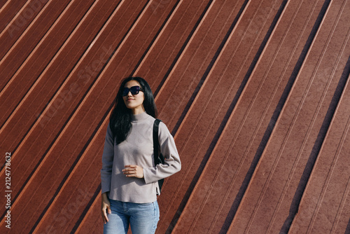 Quadro su tela Woman wearing sunglasses stands on a street against a diagonal rust wall, casual