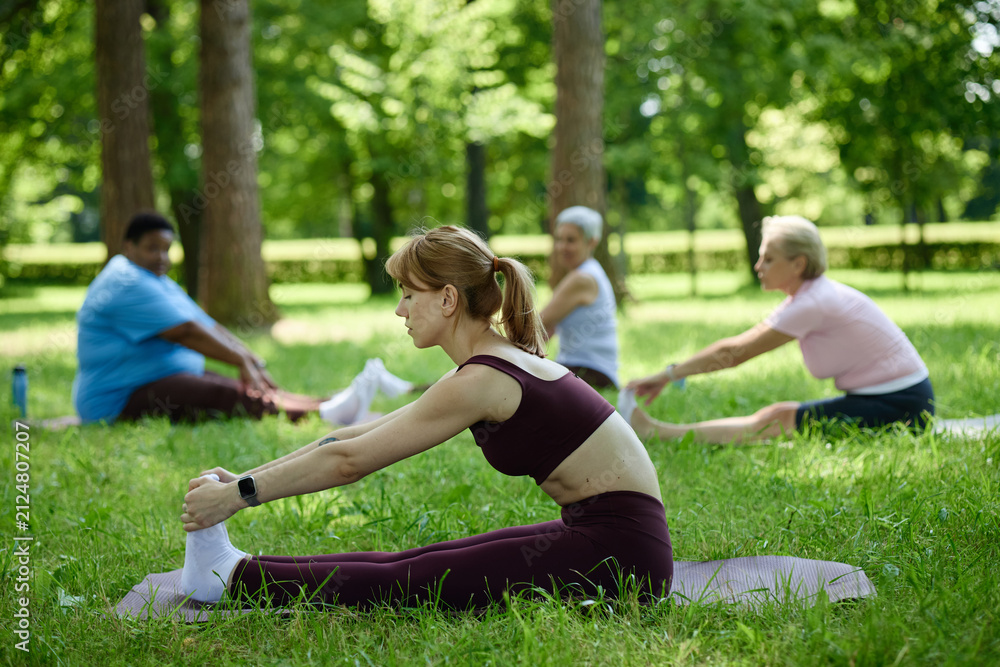 Fototapeta premium Diverse women practicing yoga in park, young Caucasian woman in foreground stretching on mat. Image represents outdoor wellness and is suitable for healthy lifestyle marketing campaigns