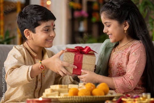 Happy Indian brother giving a beautiful gift box to his sister during Raksha Bandhan celebration. Cute siblings wearing traditional clothes with a tied rakhi bracelet and festive sweets