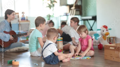 Toys are scattered on floor, children are playing with cubes and cars, teacher is playing guitar. Girl babysitter accompanies children, twang guitar during childrens play with toys activities. 