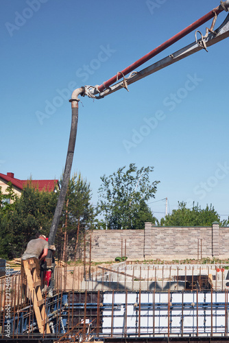 Canvas Print Vertical shot of a construction worker standing on wooden scaffolding while operating a concrete pump boom hose to pour liquid concrete into foundation formwork under a clear blue sky