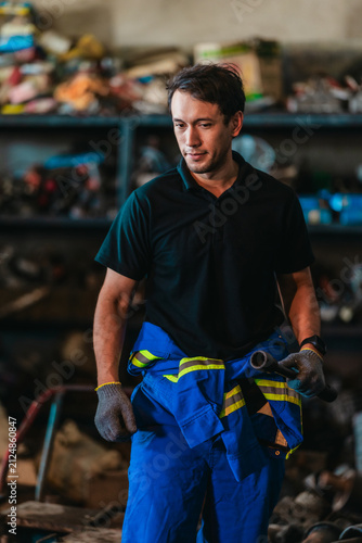 Confident male mechanic in coveralls standing in a workshop full of old machine parts