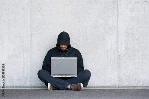 Hacker with laptop sitting in front of concrete wall