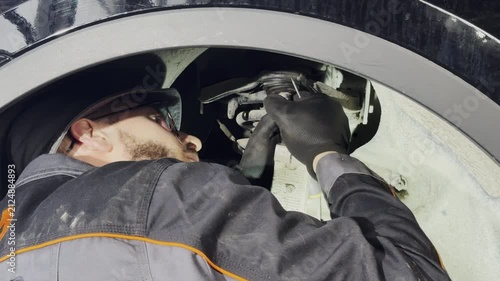 Bearded technician performs suspension repair under wheel well. Mechanic touches parts with fingers and inspects component performing task